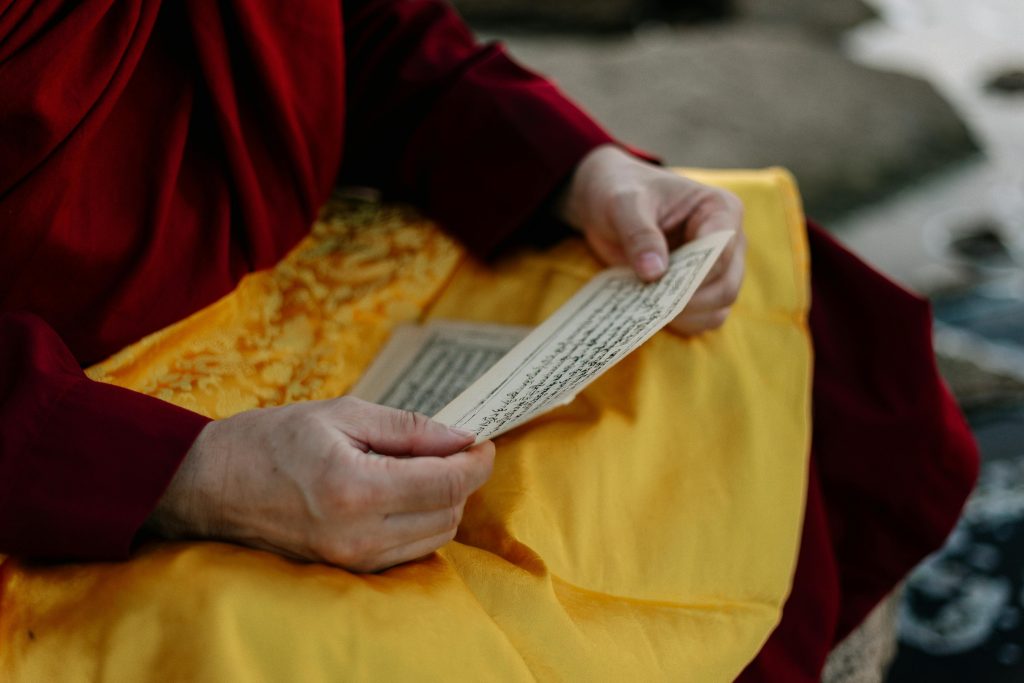 In the mantra chanting meditation, a man dressed in a red robe is deeply immersed in his practice. He sits with a yellow cloth draped over his lap, holding a mala (prayer beads) in his hand as he chants his mantra, creating a serene and focused atmosphere.