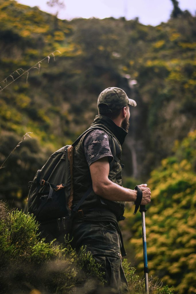 can hiking heal depression. The image shows a man in army-colored clothing standing on top of a hill, with a backpack on his back. He’s looking out over green hills, which give a sense of calm. He seems to be thinking about whether hiking can help with depression. The peaceful scenery and his thoughtful pose suggest he’s finding comfort and contemplation in the moment.