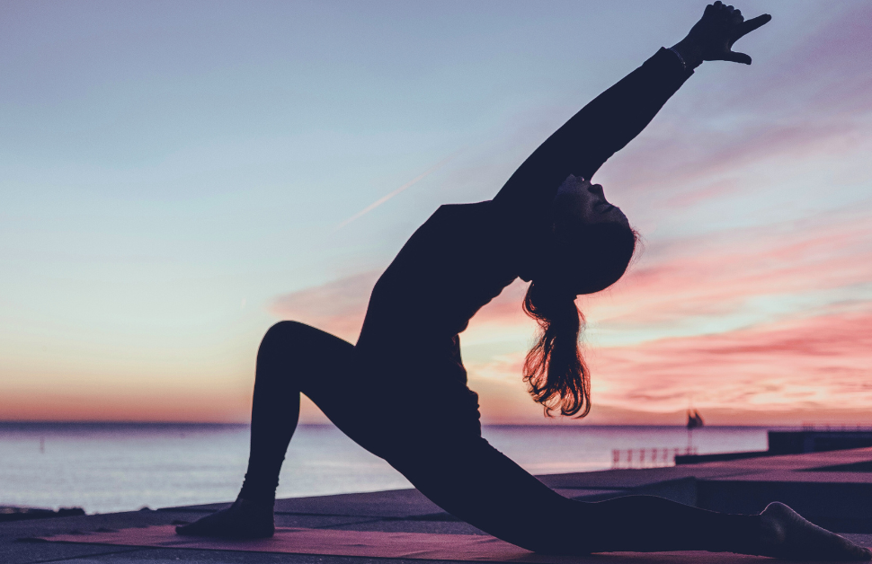 A girl practices yoga on a sandy beach, bathed in stunning sun rays with the ocean stretching out before her.