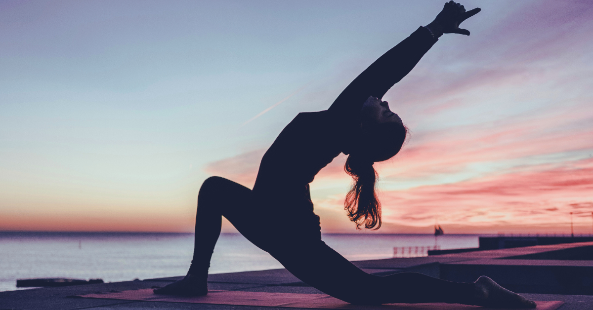 A girl practices yoga on a sandy beach, bathed in stunning sun rays with the ocean stretching out before her.