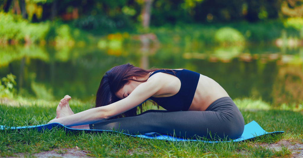 A girl is practicing yoga nidra on a mat surrounded by lush green grass and small trees, creating a tranquil oasis in nature.