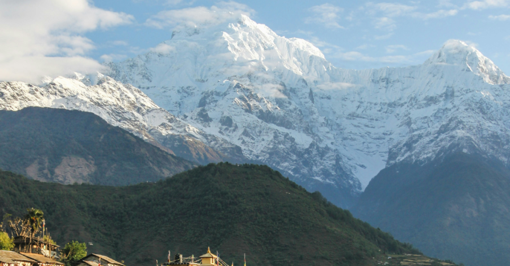 The image shows a stunning mountain view which one of the Best yoga trekking routes . In the foreground, there's a charming village nestled among green hills. The village features traditional houses and a small temple with a golden roof.
The main focus is a majestic, snow-covered mountain peak, probably Annapurna I, towering over the landscape. It’s surrounded by other high peaks, creating a dramatic and impressive scene.
The sky is a beautiful mix of blue and white with fluffy clouds, adding depth to the view. The overall feeling is peaceful and full of natural beauty.