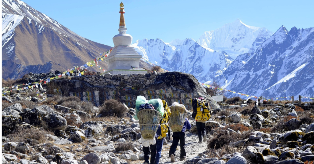 The image shows people carrying doko baskets filled with trekking supplies. They are walking along a narrow path with stones on the ground. At the top of the path, there is a small stupa adorned with hanging prayer flags. The surrounding landscape features snow-capped Himalayan peaks, adding to the breathtaking scenery.