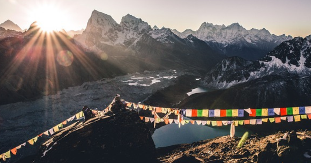 The image shows a stunning Himalayan sunrise. In the foreground, a rocky outcrop with colorful prayer flags stands out. The sun is rising behind the mountains, casting long shadows and giving the snow-covered peaks a golden glow.
The scene features a dramatic mountain range, with several peaks, possibly including Mount Everest. The mountains are surrounded by glaciers and snowfields, contrasting with the dark rocky ground.
In the distance, a calm lake, probably Gokyo Lake, reflects the mountains and sky. The overall feeling is one of peace and natural beauty.