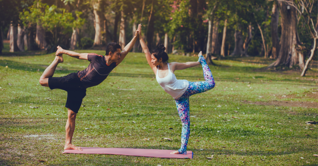 Yoga Retreat for Families: The image depicts a man and a girl standing on one leg, joining hands, on a red mat. They are surrounded by lush green grass and medium-sized trees.