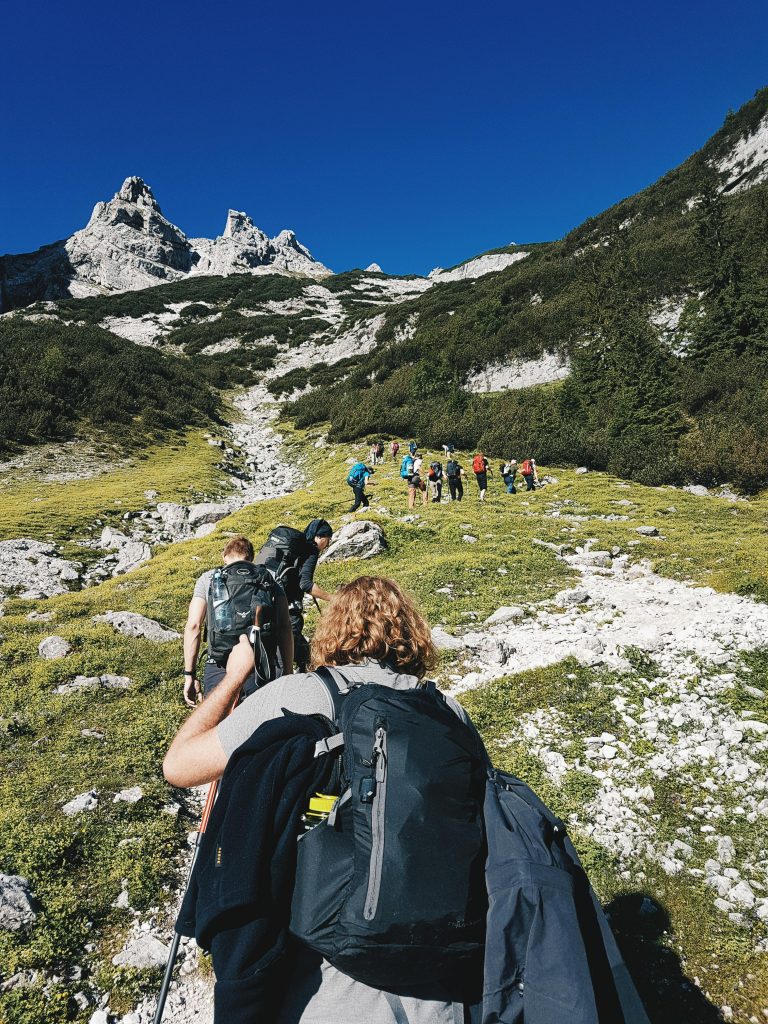 The picture depicts a safety yoga trek group trekking through a stunning landscape. They traverse a rugged terrain among lush green hills, with snow-capped mountains rising majestically in the distance. In the expansive scenery, there is a narrow crevice filled with rocks with the blue sky.