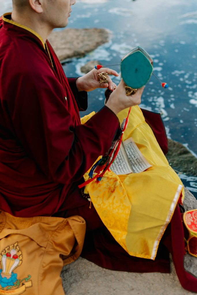 The Buddhist, dressed in red robes, is seated by the river, chanting a mantra. He holds a vajra and mala beads in his hands, with a damaru drum resting nearby. On his lap, there is a yellow cloth with the script of the mantra written above it.