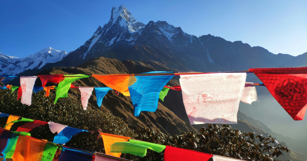 The image shows a colorful scene in the Himalayas. Prayer flags with intricate patterns and mantras flutter in the wind against the backdrop of a tall, snow-capped mountain peak, likely Mardi Himal. The bright colors of the flags contrast with the mountain and the clear blue sky. The scene reflects Himalayan culture, combining spirituality with beautiful natural scenery.