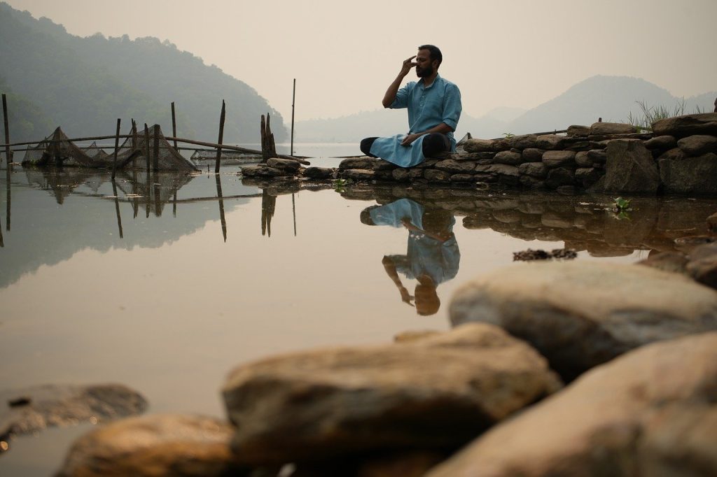 The image shows a man practicing breathing exercises (pranayama) on a rock by a peaceful pond. The background features hills and a clear blue sky reflected in the water. It looks like a calm and beautiful place to do yoga - Integrating Pranayama Techniques into Your Yoga Trek 