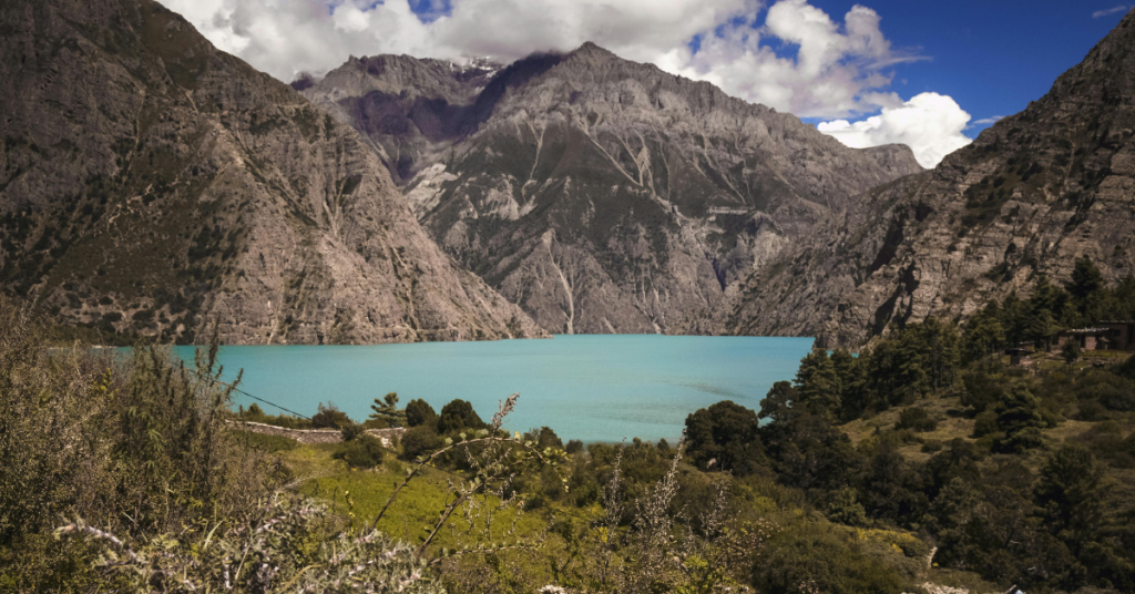 The image shows a beautiful mountain lake set in a dramatic landscape. In the foreground, green plants and a small village line the shore, offering a peaceful contrast to the rugged mountains.

The lake is a clear turquoise, reflecting the surrounding mountains and sky like a mirror. Tall, rocky cliffs rise sharply around the lake. The sky above is filled with fluffy white clouds, adding depth to the scene. Overall, the atmosphere is calm and picturesque. 