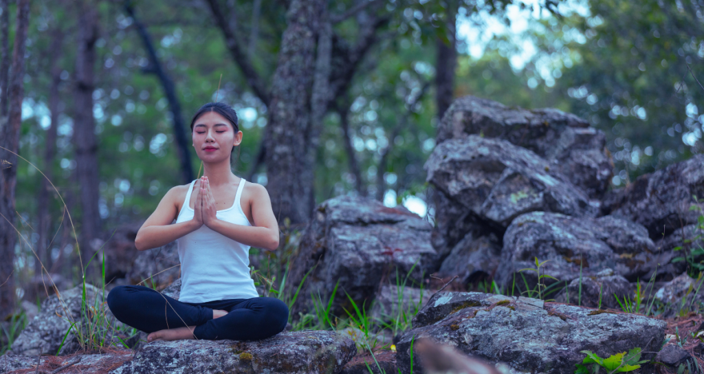 The image depicts a girl practicing yoga on a trek through a forest, surrounded by stones and towering trees. She sits on a stone, embodying a digital detox as she connects with nature in this serene, natural setting.