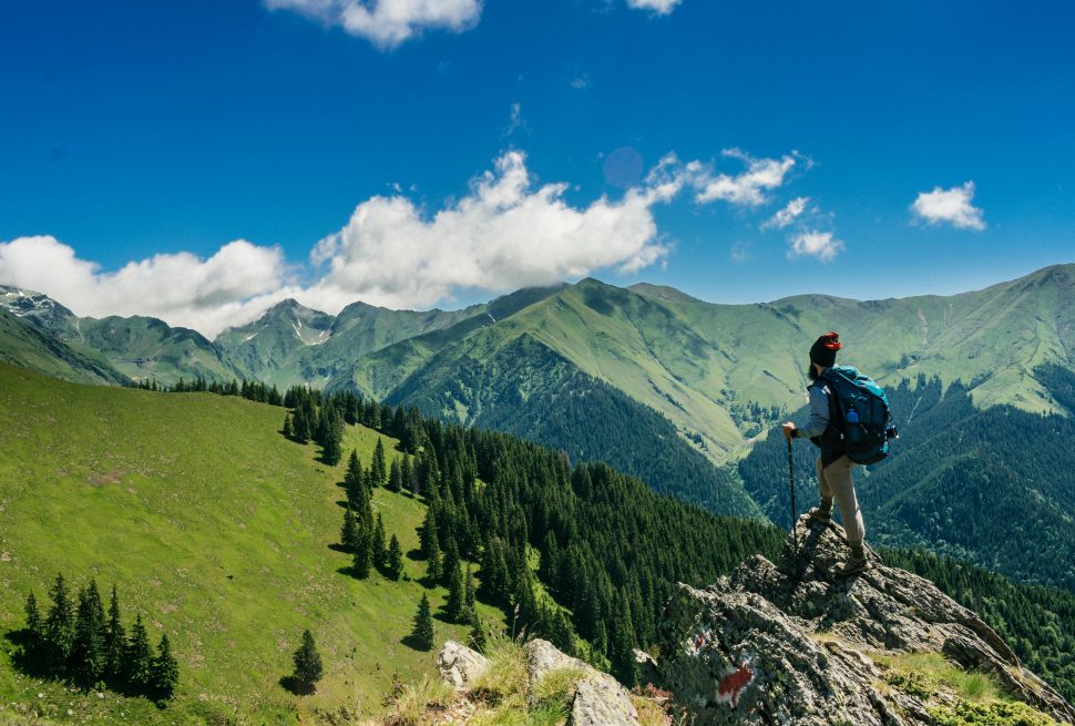 Under the blue and cloudy sky there is green hills and on the left side of the picture the hills contain the few green trees and on the right hand side there is rock where the man is standing with the climbing stick and bag