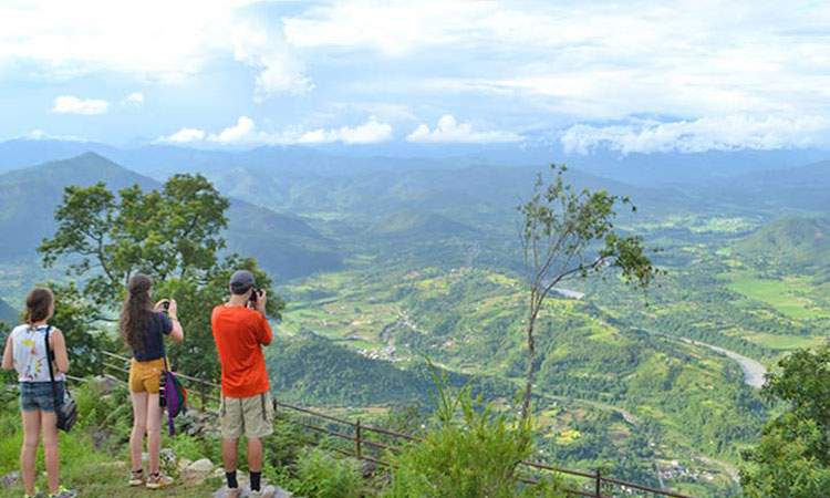 The image shows a view from Champadevi, where two girls and a man are taking photos of the beautiful green landscape and lakes visible from the top. They are surrounded by green hills under a clear blue sky.