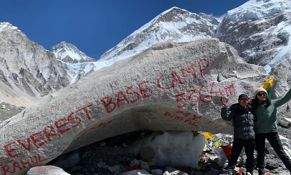 Two men stand beside a rock on Everest, which is marked with "Everest Base Camp 5364m." Behind the rock, Mount Everest rises majestically under a clear blue sky.