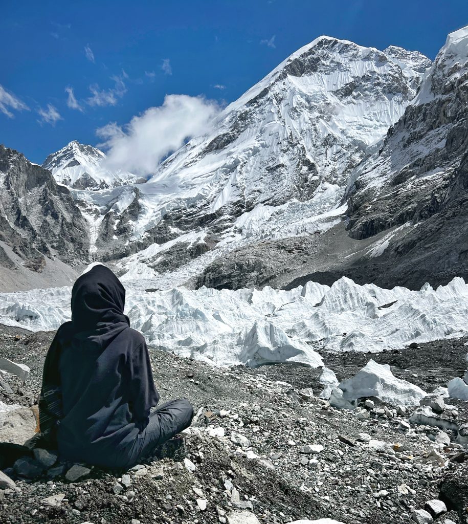 this is the picture describe that one man s doing yoga at the base of the Everest  where the himal is covered with the snow and clouds on the blue sky and the man is sitting ont he rock and small pebbles