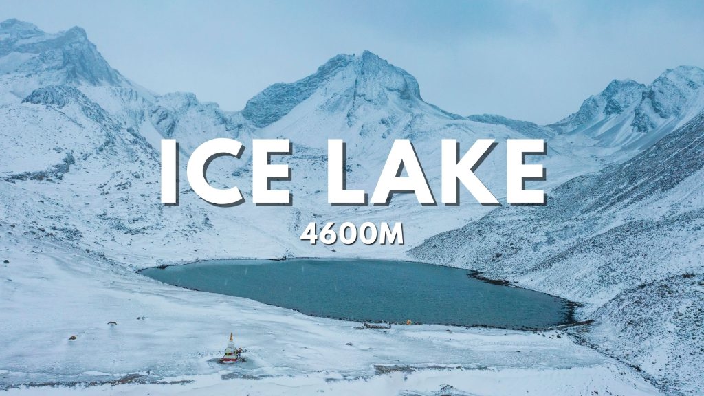 The image shows a frozen lake surrounded by snow-capped mountains. The lake is a deep blue color, and the mountains are a stark white. A small building or shrine is visible on the shore of the lake.