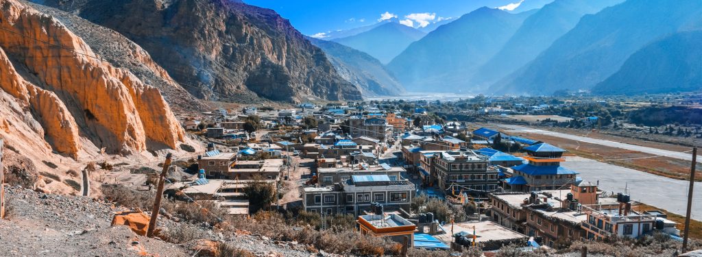 The image showcases a breathtaking view of the town of Jomsom, nestled amidst the majestic Annapurna mountain range. The town is perched on a hill, with traditional stone buildings and colorful rooftops dotting the landscape. A small airport is visible in the distance, nestled between the mountains. The towering peaks of the Annapurna massif provide a dramatic backdrop, with their snow-covered summits contrasting against the blue sky.








