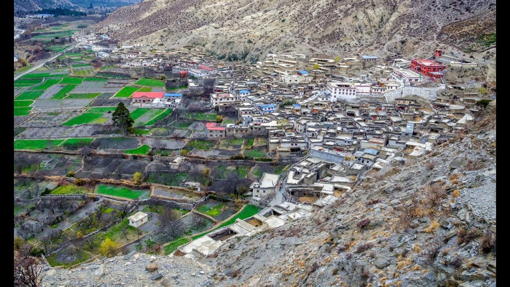 The image depicts a picturesque Himalayan village nestled amidst a rugged mountain landscape. The village is characterized by traditional stone houses with flat roofs, arranged in a terraced pattern along the hillside. Green fields and terraced agriculture can be seen surrounding the village. The mountains in the background are majestic and snow-capped, creating a stunning natural backdrop.