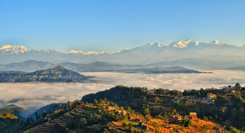 Under the blue sky, there are the Himalayas, with green hills below them surrounded by clouds. In front of the cloudy hills, a village is visible, along with terrace farming.