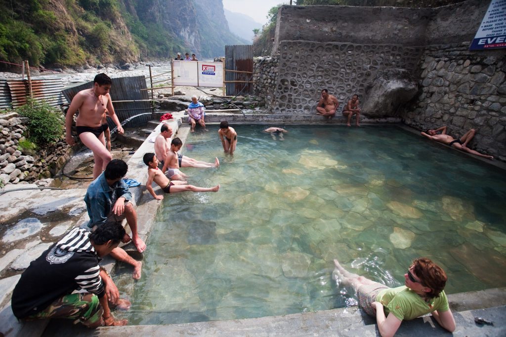 There is a blue Tatopani-like pond where many people are enjoying the serene waters. Beside the pond, a large stone rests on the shore, and in the background, a lush green hill rises majestically.