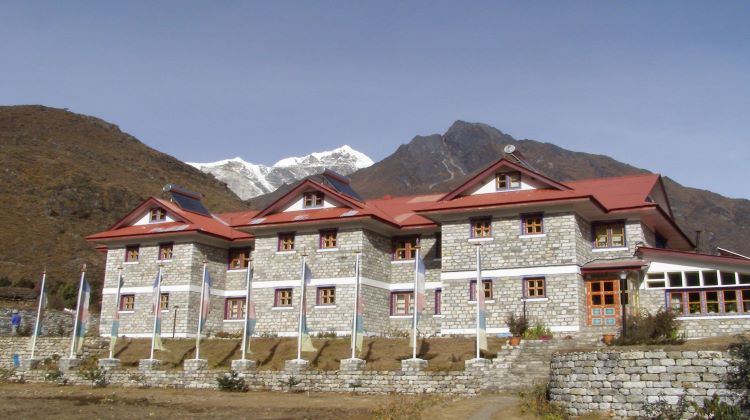 The lodge and accommodation are depicted with colorful flags fluttering behind it. The setting includes lush green hills and the towering Himalayas in the background, creating a picturesque and inviting atmosphere.