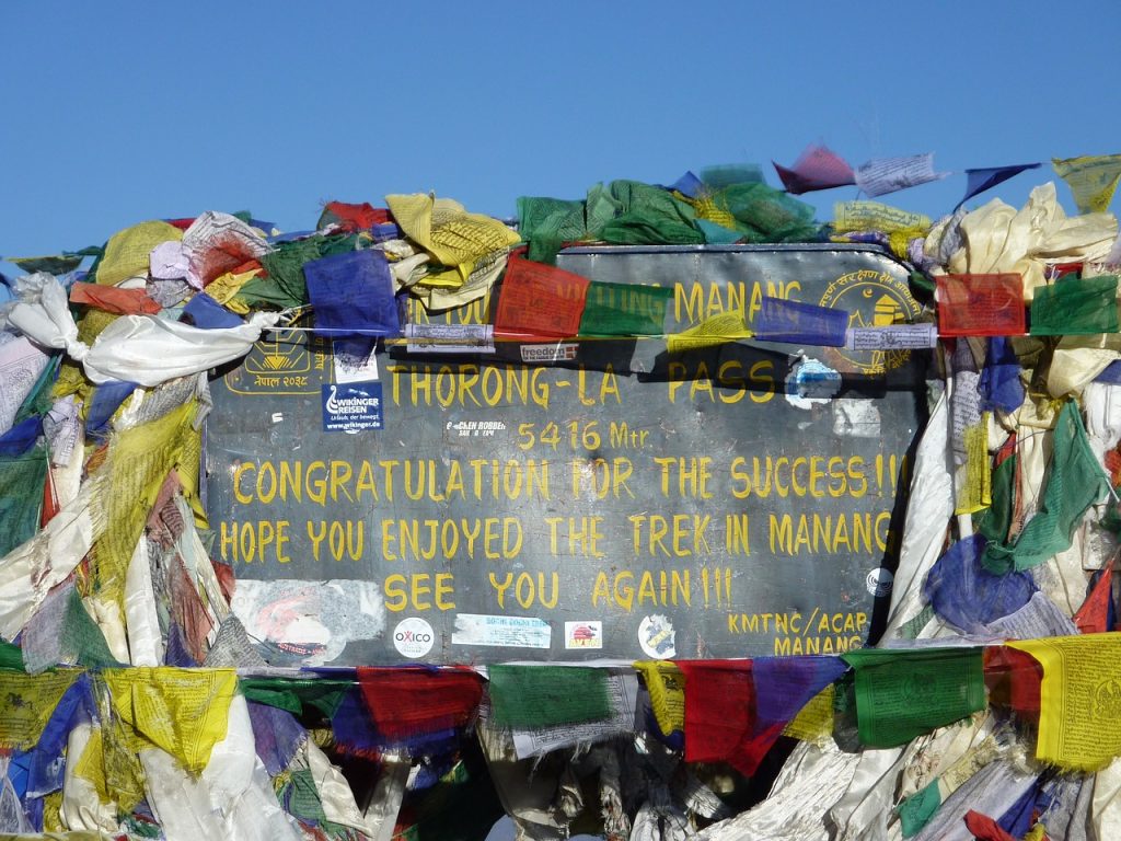 The image shows a signpost at Thorong La Pass, a high mountain pass in Nepal. The sign congratulates visitors on reaching the pass and expresses hope that they enjoyed their trek in Manang. It also includes the elevation of the pass (5416 meters) and the names of the organizations responsible for the sign (KMTNC/ACAP Manang). The sign is surrounded by a colorful array of prayer flags.