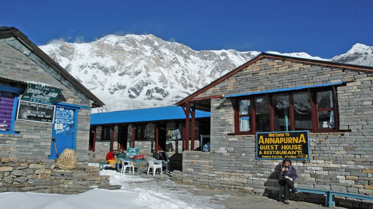 The picture shows a man seated and enjoying tea at the Annapurna Guest House and Restaurant. The hotel, prominently labeled, is situated with a stunning mountain towering above it.