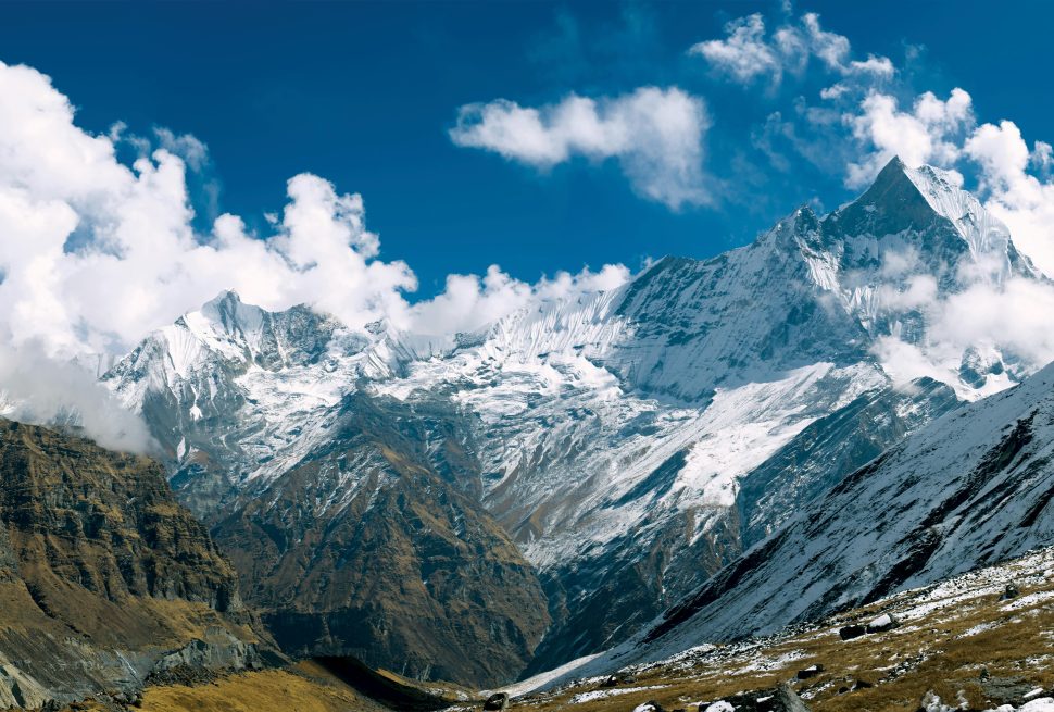 The image showcases a breathtaking panoramic view of the Hidden Gems of Annapurna Circuit. Towering peaks, including Annapurna I and Annapurna South, are adorned with snow and glaciers, contrasting against the clear blue sky. Dramatic clouds drift across the mountaintops, adding depth and texture to the scene. Lush green valleys and rugged terrain complete the picturesque vista, revealing the hidden treasures of this iconic trekking route.