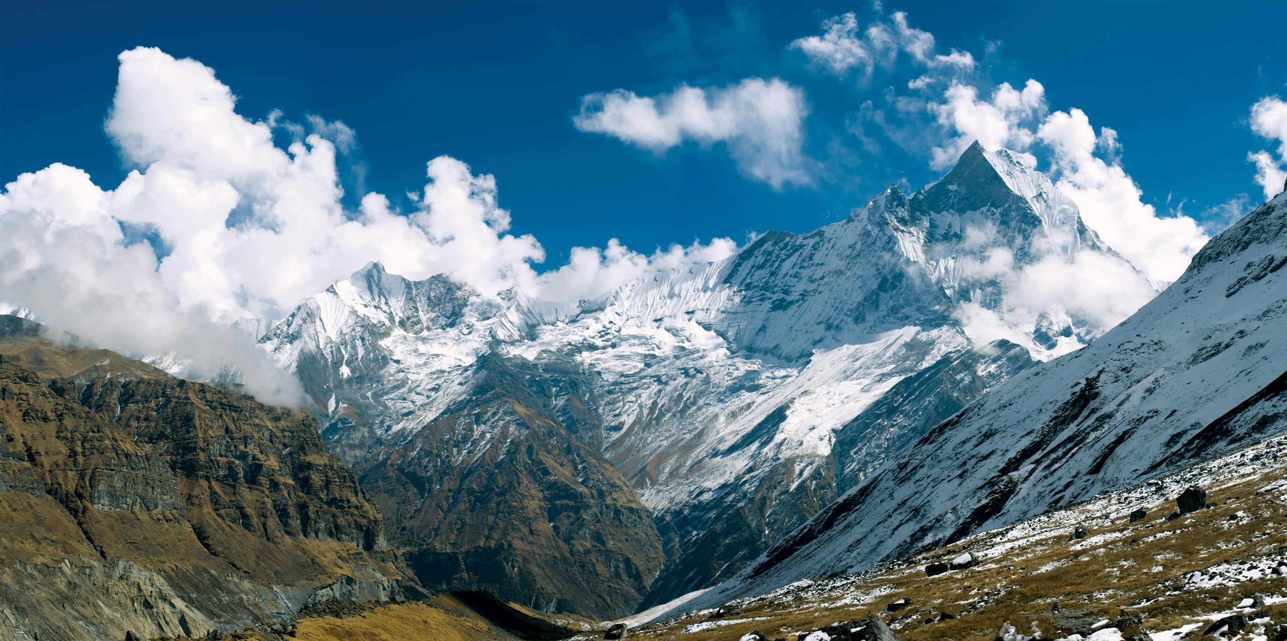 The image showcases a breathtaking panoramic view of the Hidden Gems of Annapurna Circuit. Towering peaks, including Annapurna I and Annapurna South, are adorned with snow and glaciers, contrasting against the clear blue sky. Dramatic clouds drift across the mountaintops, adding depth and texture to the scene. Lush green valleys and rugged terrain complete the picturesque vista, revealing the hidden treasures of this iconic trekking route.