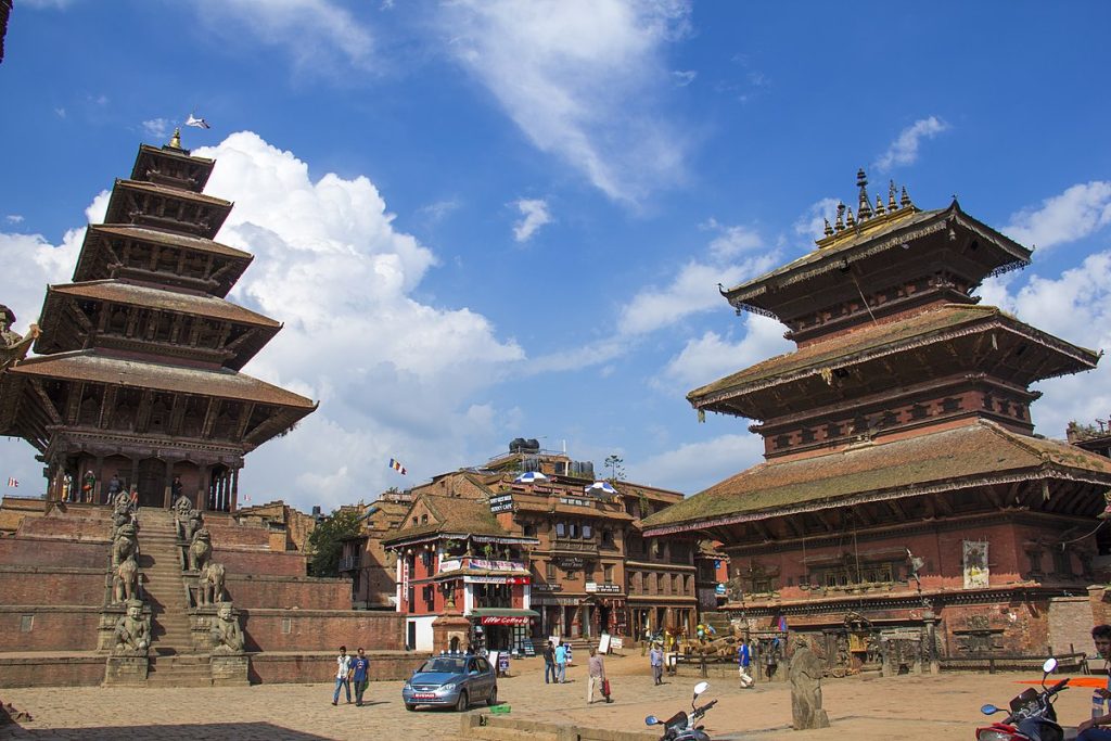 The picture shows two tall, wooden temples with many levels. There are people walking around the square, and there are cars and motorcycles. The sky is blue with white clouds.