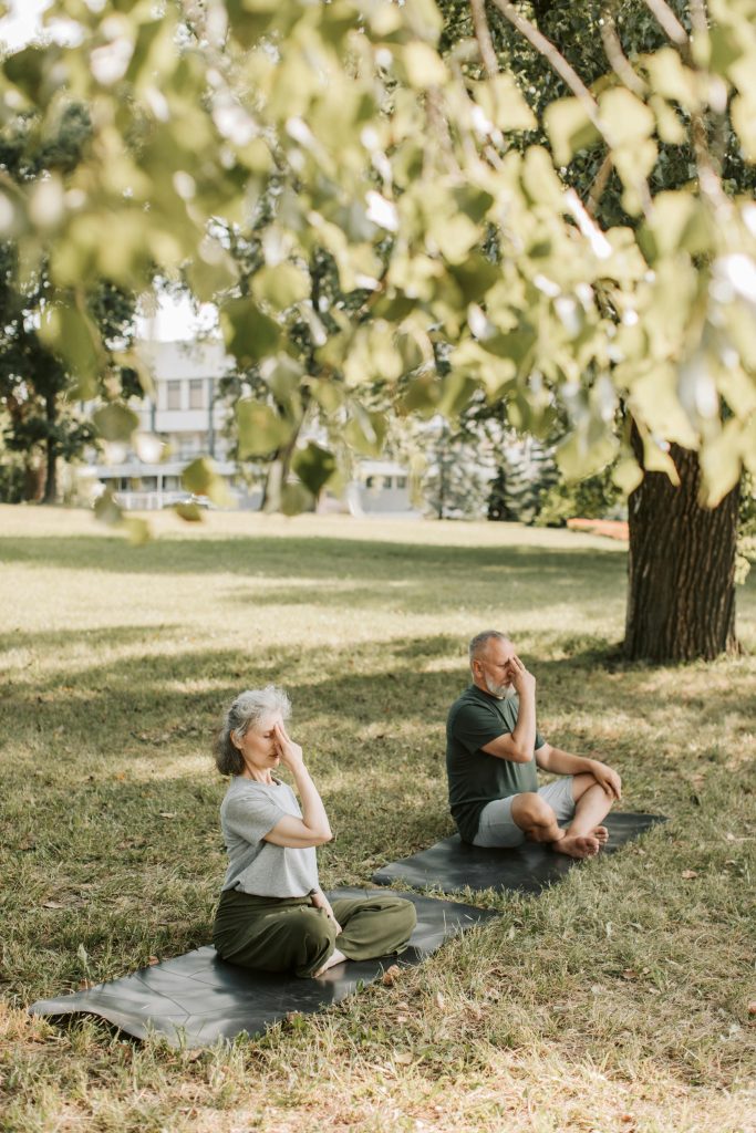 Two men sit peacefully in a serene environment, with a house and trees behind them and dry grass covering the land around them.