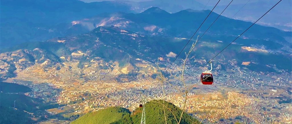 The image shows a stunning view from above a mountainous area. In the foreground, a red cable car is hanging from thick cables, moving over a busy city below. The city is full of buildings and roads that stretch out far. Beyond the city, there are rolling hills and mountains that go on towards the horizon, lit up by the setting sun. The sky has beautiful shades of blue, pink, and orange, making the scene even more gorgeous.