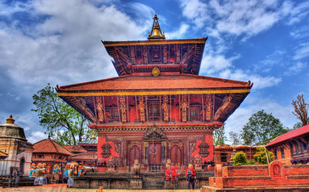 under the blue sky the picture is of "Changunararayan" and aside the main temple ther is small other temples as well and different people are walking around the temple 