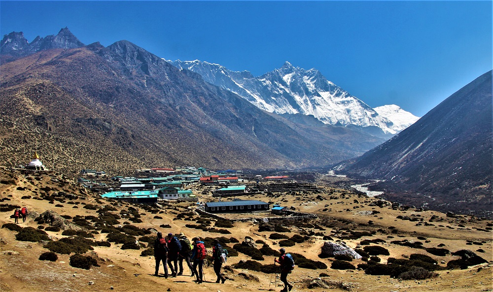 The village is bathed in blue sky, featuring a variety of low-density, colorful houses. People are trekking towards the mountain, which rises prominently behind the hills. Small bushes are scattered throughout the landscape, adding to the picturesque scene.