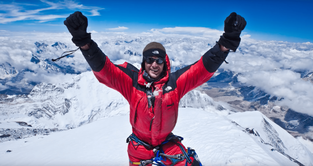 The picture shows a man standing on the summit of Everest, with a mix of cloudy and blue sky above and the mountain’s snowy peak surrounding him.