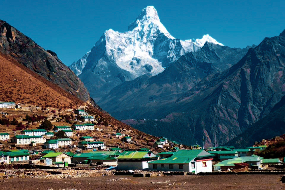 This is a view of Everest Base Camp, where lodges and hotels are situated at the base of the towering Everest. The houses are nestled at the foot of verdant green hills, which are majestically dominated by the towering presence of Everest.