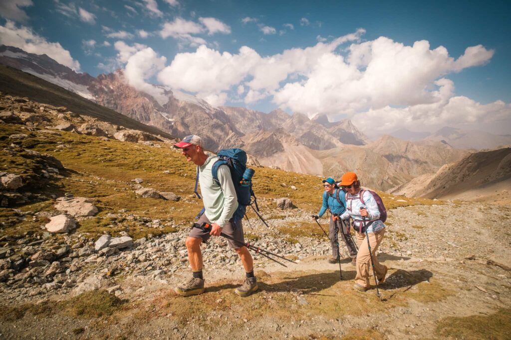 Three men are trekking, each carrying a stick and a backpack. They navigate through rocky terrain and green fields. Above them, the blue sky is dotted with clouds, and beneath the clouds, rolling hills stretch out in the distance.