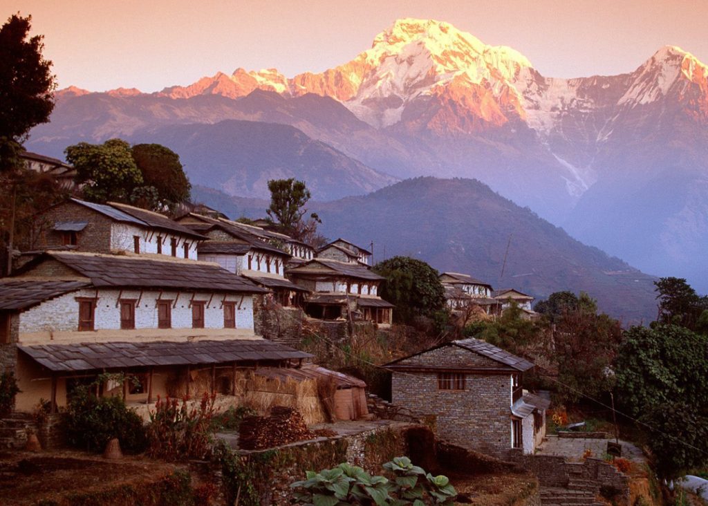 The image showcases a picturesque Himalayan village nestled amidst a breathtaking mountain landscape. The village is composed of traditional stone houses with slate roofs, arranged in a terraced pattern along the hillside. A majestic snow-capped peak dominates the background, bathed in the warm glow of the setting sun. Lush greenery, including trees and terraced fields, surrounds the village, creating a harmonious blend of human settlement and natural beauty.