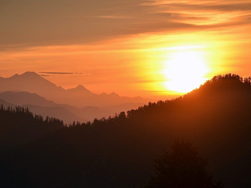 The image shows a stunning sunset over mountains. The sky is filled with bright orange and pink colors, casting a warm light on the dark hills and forests. The sun is partly hidden by clouds, creating a dramatic and magical look. The distant mountains appear soft and dreamy, adding to the peaceful and awe-inspiring atmosphere.