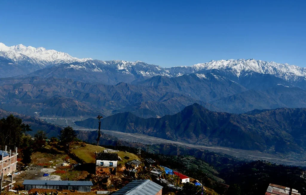 The image shows a beautiful mountain landscape. In the foreground, there is a small village with houses made of brick and stone. The mountains in the background are covered in snow and look very tall. The sky is blue and clear.