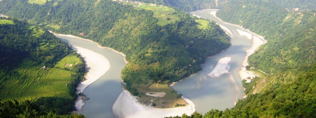 The image showcases a breathtaking aerial view of the Kali Gandaki River winding through a lush Himalayan valley. The river's meandering path creates intricate loops and bends, surrounded by verdant hillsides dotted with terraced fields and small settlements. The water appears to be a serene blue-green color, contrasting against the earthy tones of the surrounding landscape. The overall scene is one of natural beauty and tranquility.