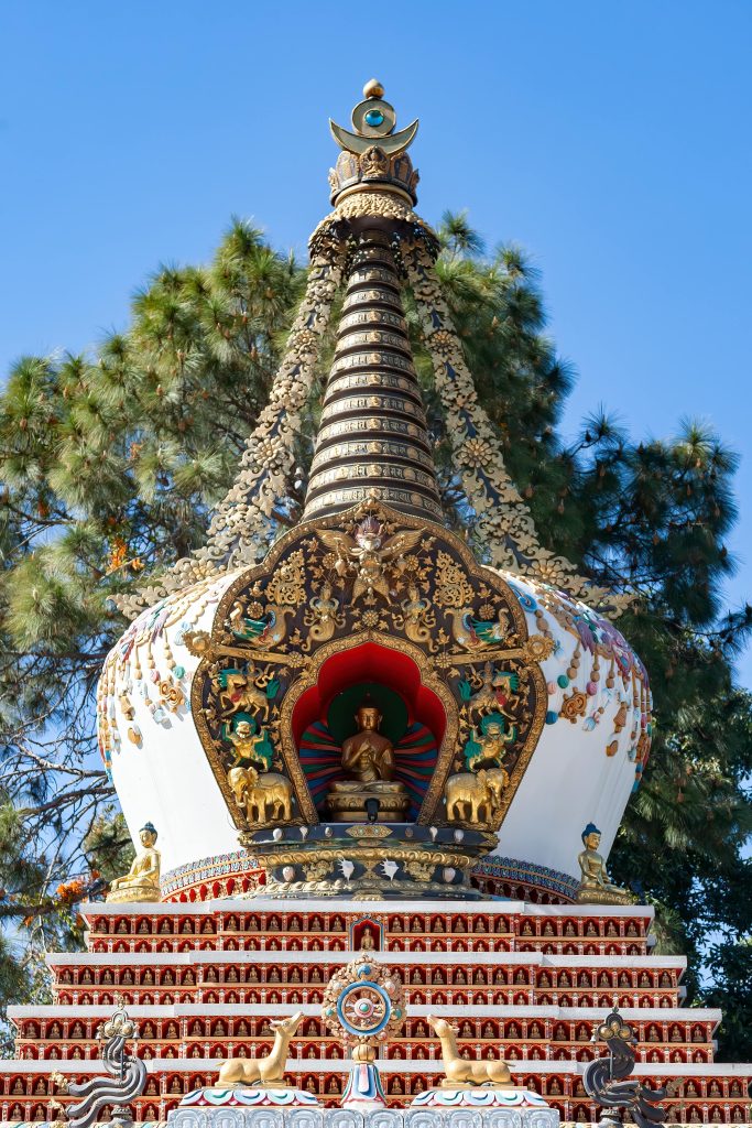 The image shows a stunning stupa, a type of Buddhist structure. It's beautifully decorated with detailed carvings, covered in gold, and painted in bright colors. In the center, there’s a figure, probably the Buddha, sitting in a red-framed space. Around the stupa, there are smaller figures and symbols, highlighting its religious importance. The clear blue sky makes the monument stand out even more.