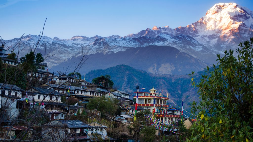 A monastery and various houses are nestled together, set against a backdrop of green hills beneath a white mountain. The sun shines brightly on the mountain under a clear blue sky.