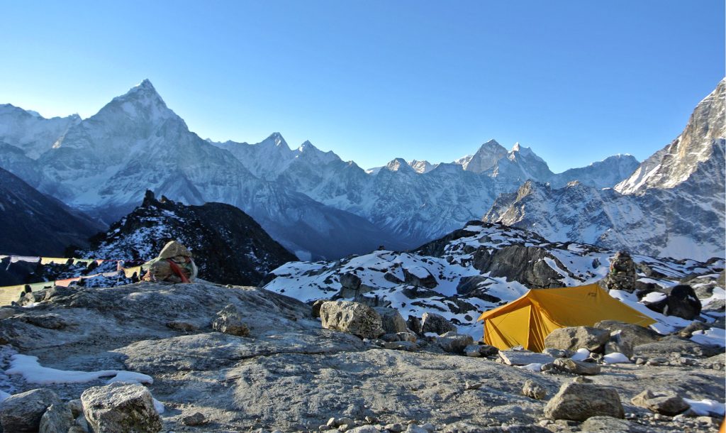 A tent is nestled between various stones and rocks at the base of the Himalayas, surrounded by towering peaks under a clear blue sky.