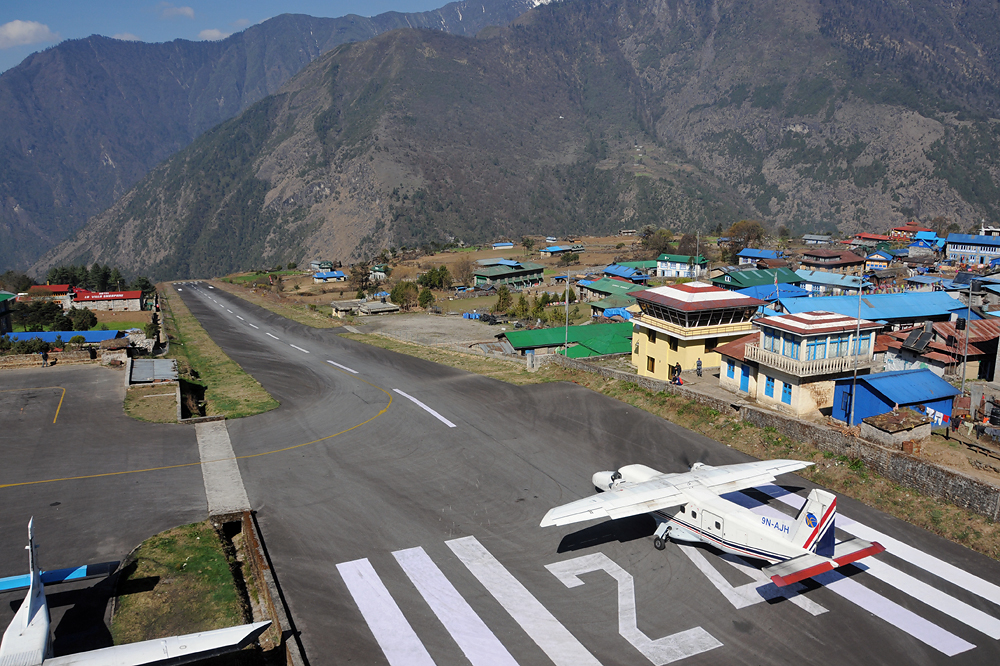 This is Lukla Airport, known as the most dangerous airport in Nepal. The scene features an airplane on the runway, surrounded by various houses. In the background, green hills stretch out beneath a clear blue sky.