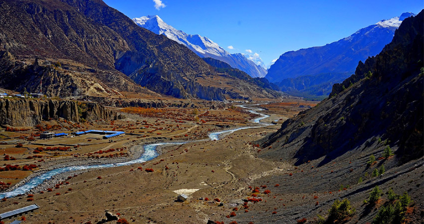 The image depicts a breathtaking mountain valley with a winding river flowing through it. The valley is surrounded by towering, snow-capped mountains, and the sky is a clear blue. There are a few small settlements visible along the river, and some terraced fields can be seen on the slopes of the mountains. The overall atmosphere of the image is one of peace and tranquility.