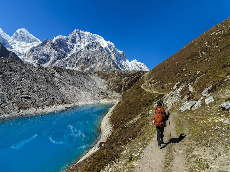 The scene features a snow-capped mountain with a blue pond below it. A trekker is walking along a narrow path beside the pond, with lush green hills flanking the trail.