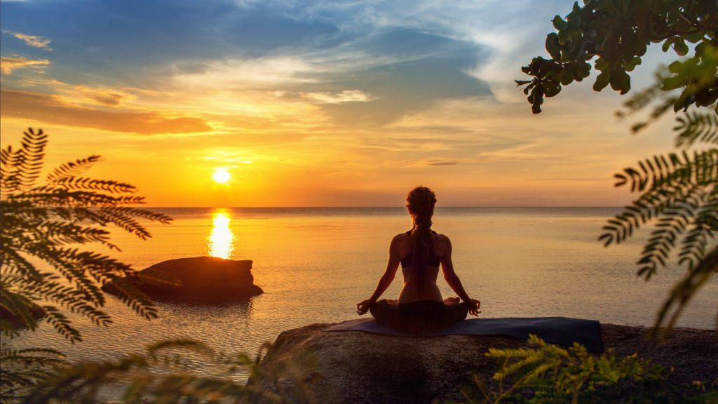 The girl is doing yoga on a rock with her mat facing the setting sun. In front of her, there’s the ocean, and to the left on the rock, there are some leaves behind her.