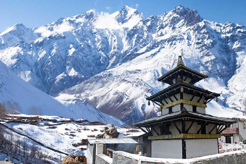 The image depicts a serene temple nestled amidst a breathtaking Himalayan landscape. A snow-capped mountain range towers over the temple, creating a dramatic backdrop. The temple itself is a traditional Nepalese structure with a tiered roof and intricate carvings. The sky is a clear blue, and the sun casts a warm glow over the scene.