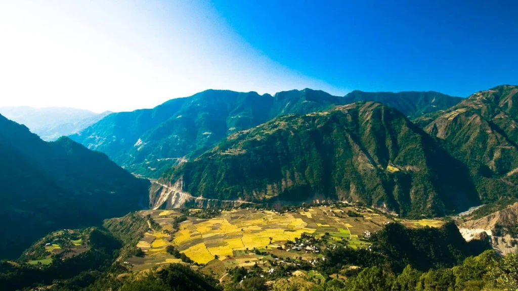 The picture shows a beautiful mountain scene. The sky is blue and clear. The mountains are green and tall, with deep valleys between them. There is a small village with fields at the bottom of the mountains. It looks peaceful and natural. The mountains are very tall and steep, with lots of rocks and trees. The fields are green and flat, and the village has many small houses.